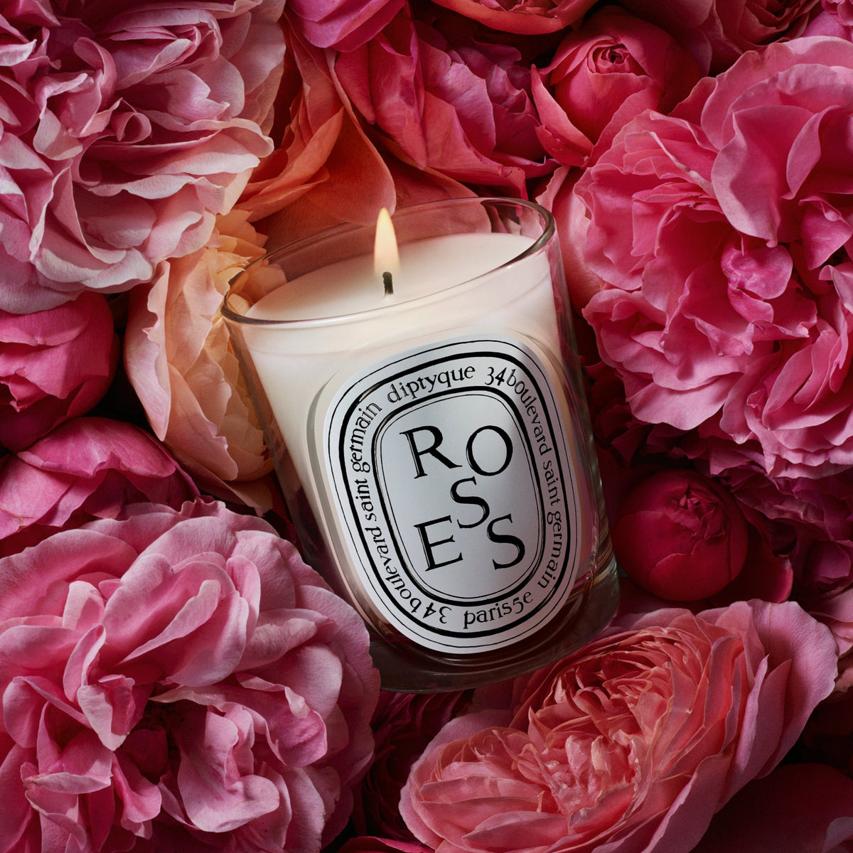 Diptyque Roses candle burning in a bed of fresh cut roses, one of the best rose candles that is not sweet