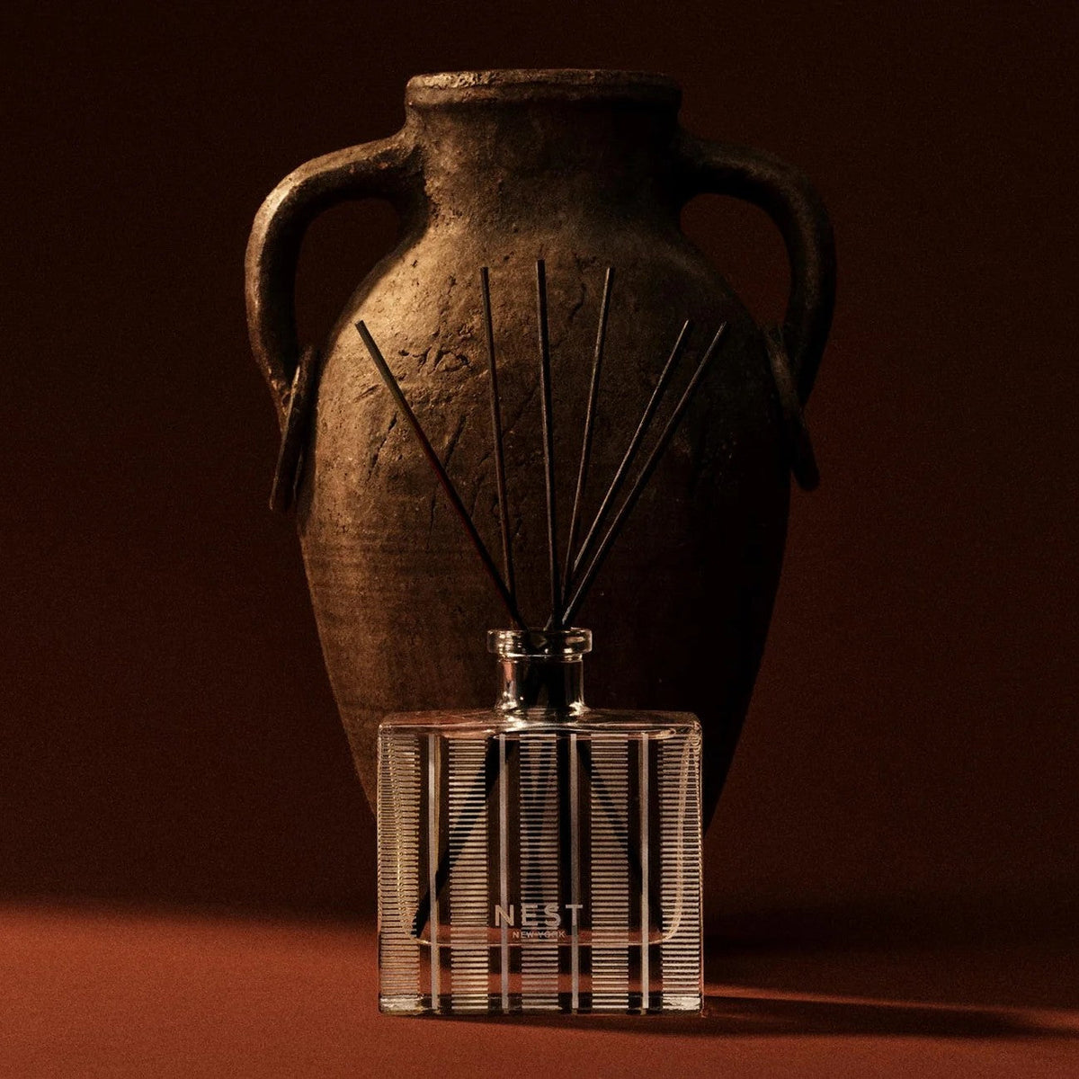 Nest Moroccan Amber reed diffuser beside a dark clay amphora in warm low light: the entryway scent that makes a house smell good like a boutique hotel