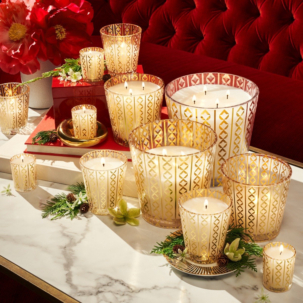 Nest Holiday candles in gold-patterned glass burning on a marble table with red peonies and pine, the best Christmas candles of 2026 tested by scent family