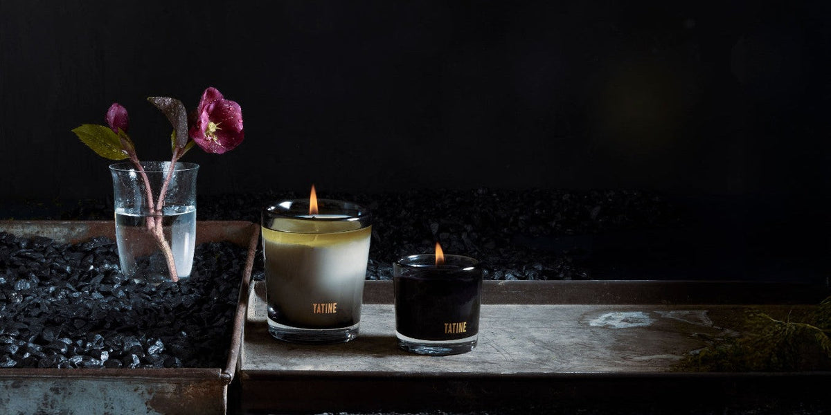 Lit Tatine amber glass Classic candle and black wax Petite on a rusted tray with black gravel and a hellebore stem