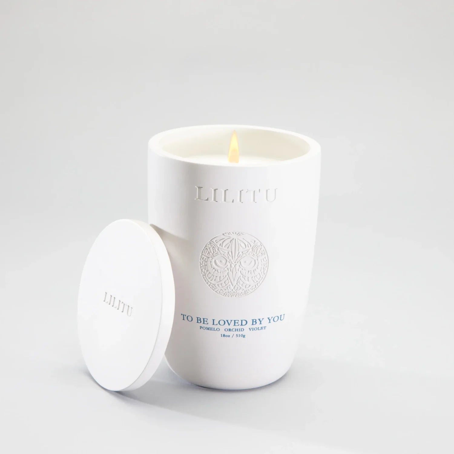 Lilitu To Be Loved By You white ceramic candle, lit, with its lid resting against the vessel