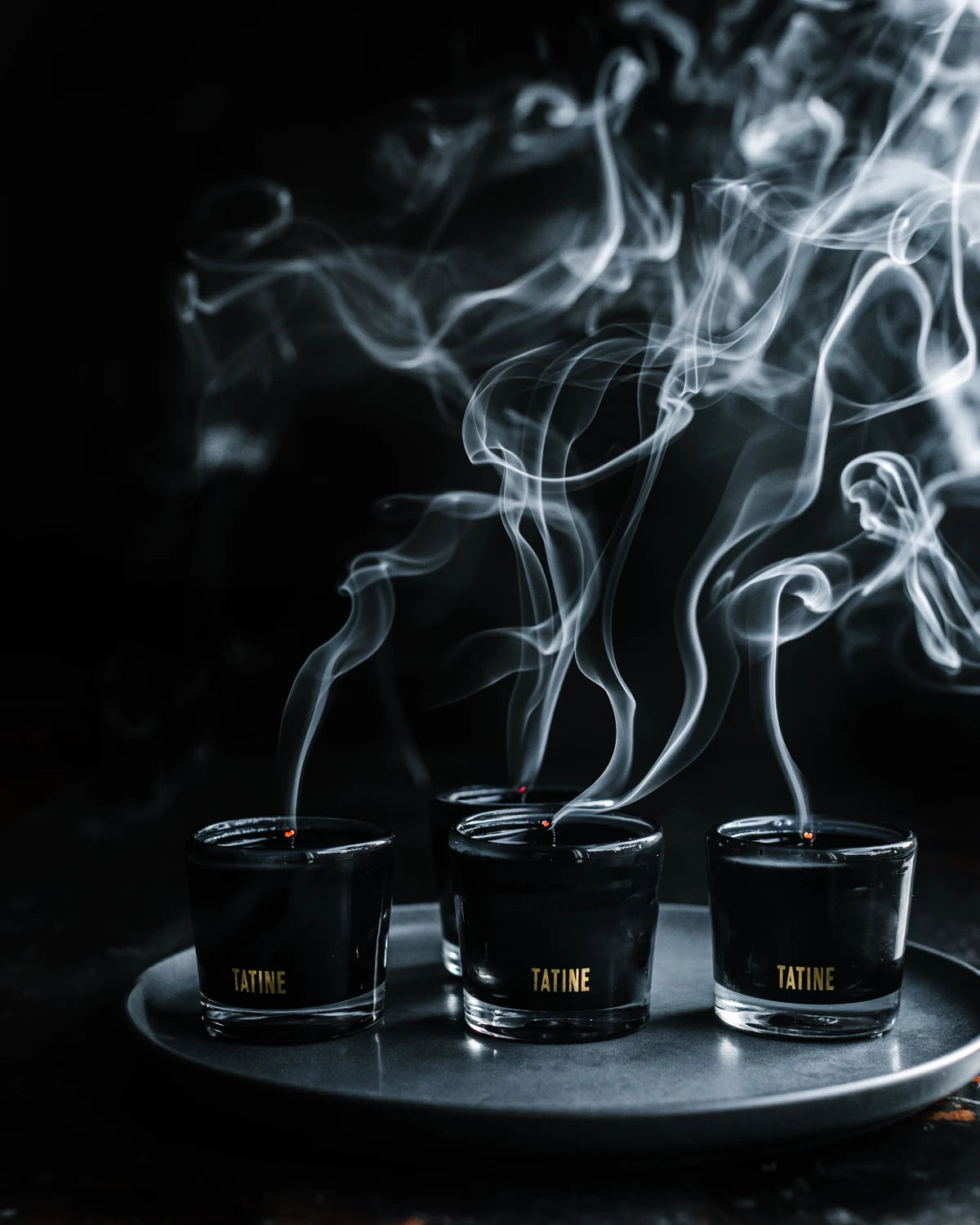 Tatine black wax candles just blown out, smoke trails rising against a dark background