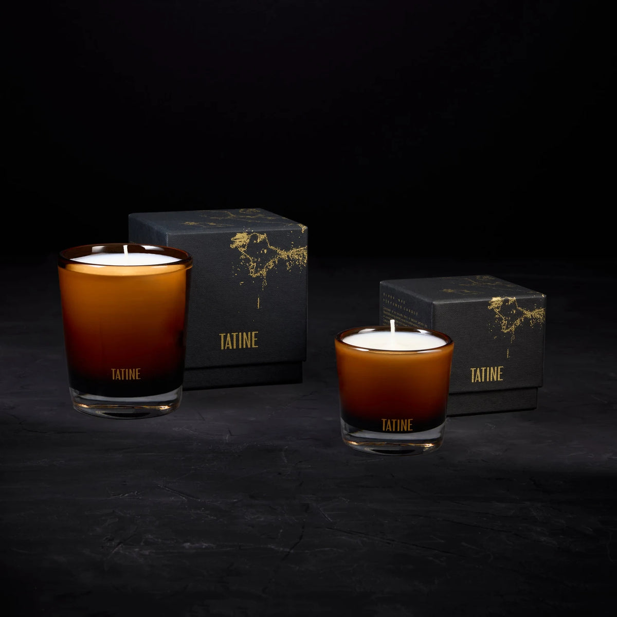 Tatine amber glass Classic and Petite candles lit beside gold-splattered black gift boxes